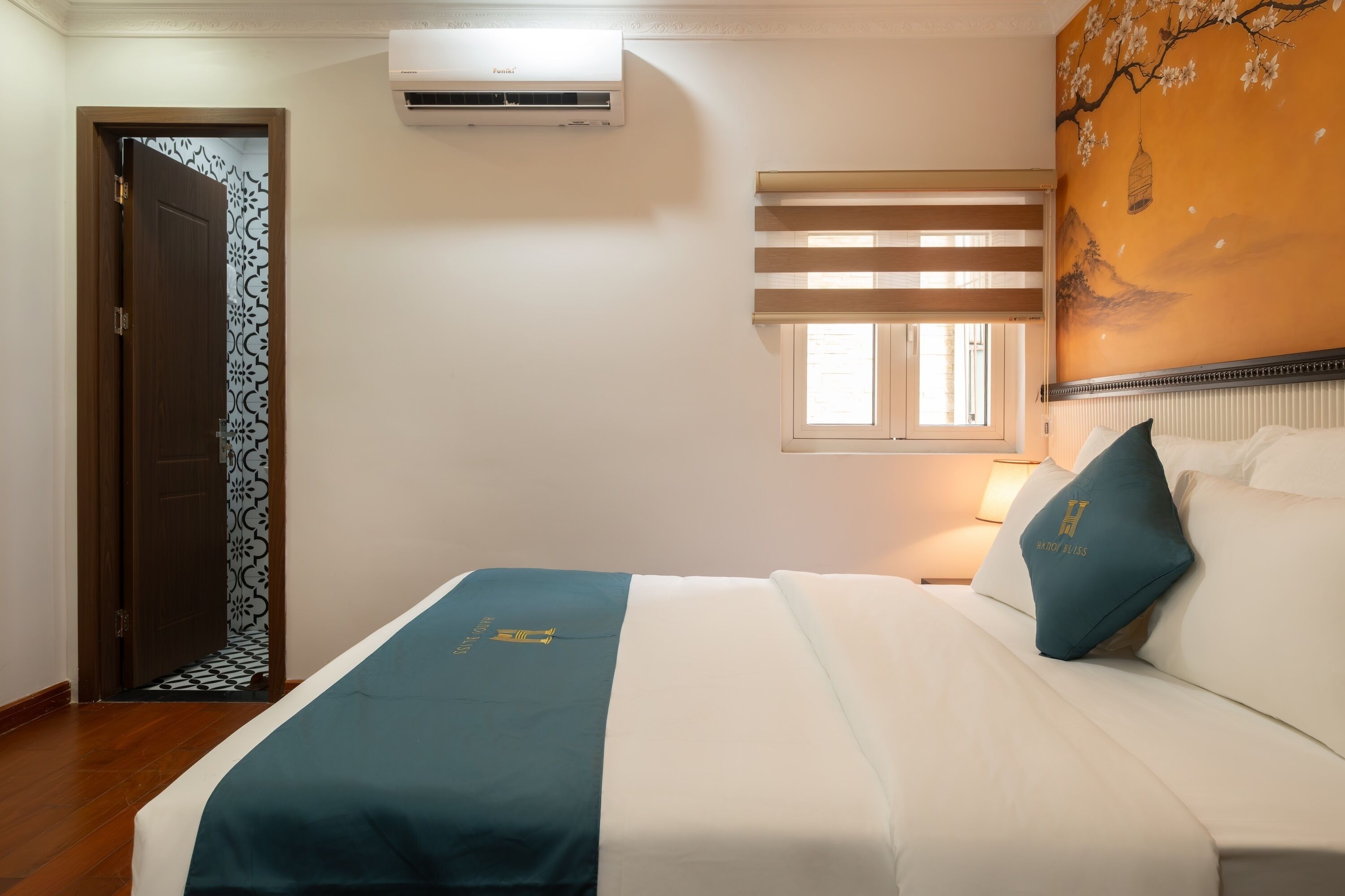 Habitación de hotel con una cama, una ventana con persianas, un equipo de aire acondicionado, una puerta con diseño decorativo y una pared con un mural floral.