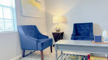 A room with two blue armchairs, a small table, a lamp, and a bookshelf with books.