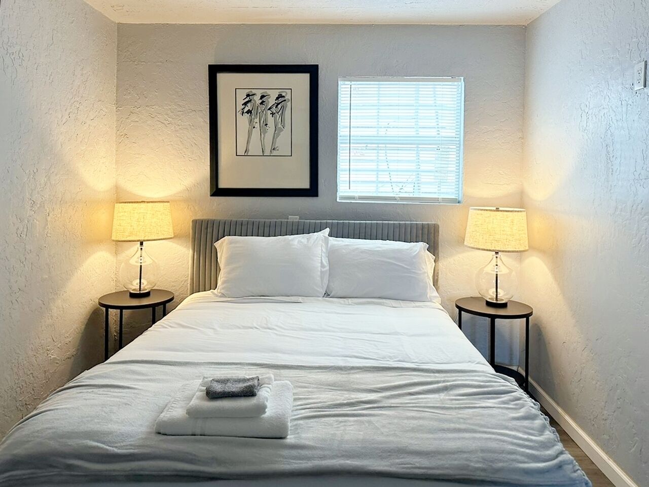 A neatly made bed with white linens, two bedside lamps, a framed artwork above the bed, and a window with blinds.