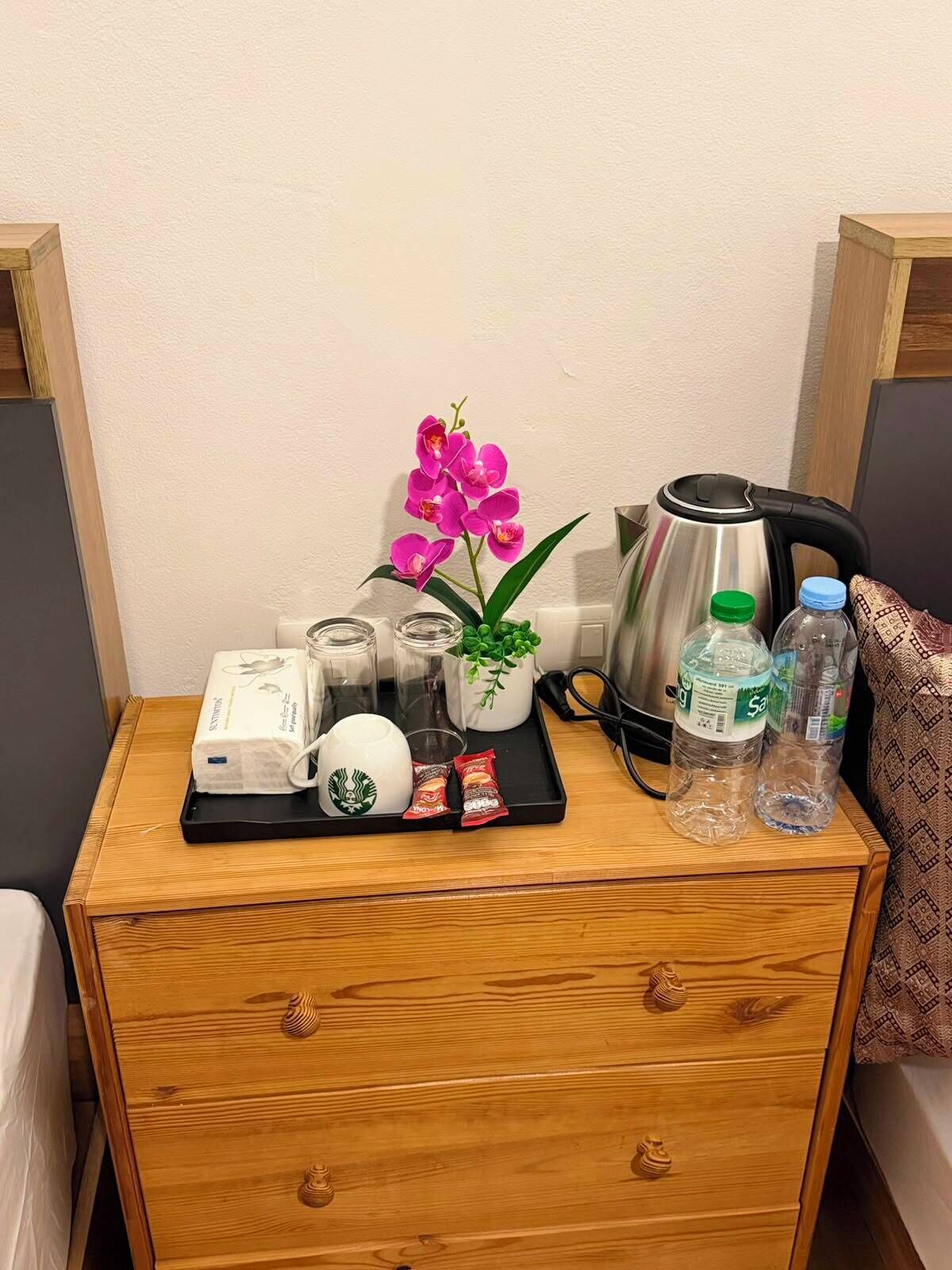 Una mesita de noche de madera con una bandeja que contiene un vaso de Starbucks, una planta, una tetera y agua embotellada.