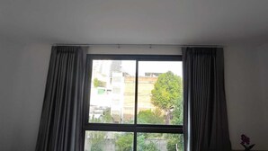 Una habitación con un ventanal, cortinas y vistas a edificios y árboles.