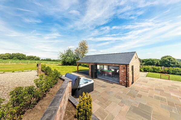 A brick-built outdoor pool area with a hot tub, a covered seating area, and a view of a green field. - Fairvale House (Northwich)