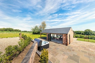 A brick-built outdoor pool area with a hot tub, a covered seating area, and a view of a green field.