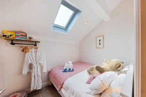 Un dormitorio con tragaluz, una cama con colcha rosa, un osito de peluche y una estantería con libros y un reloj.