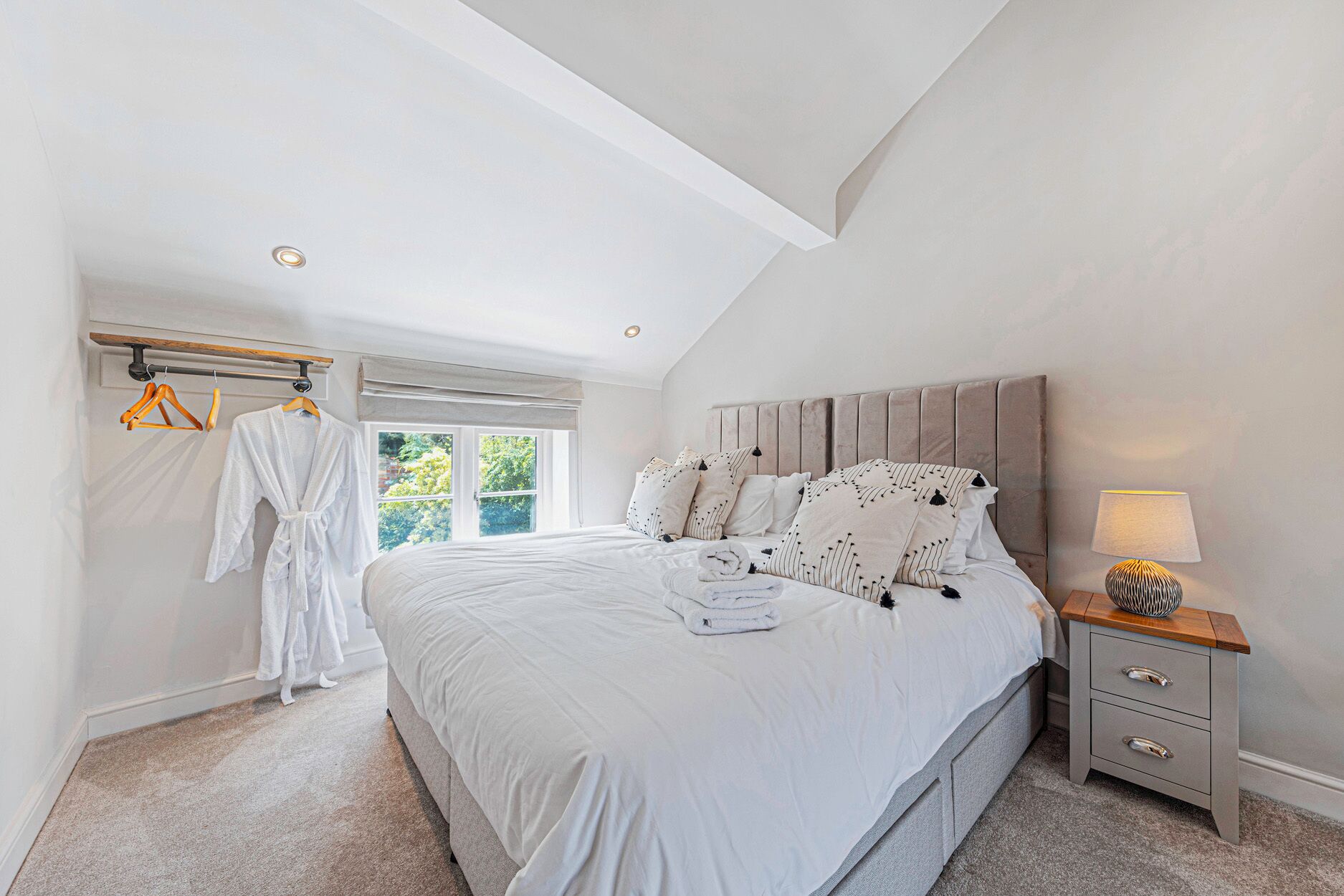 A bedroom with a large bed, a bedside table with a lamp, and a window with a view of greenery.