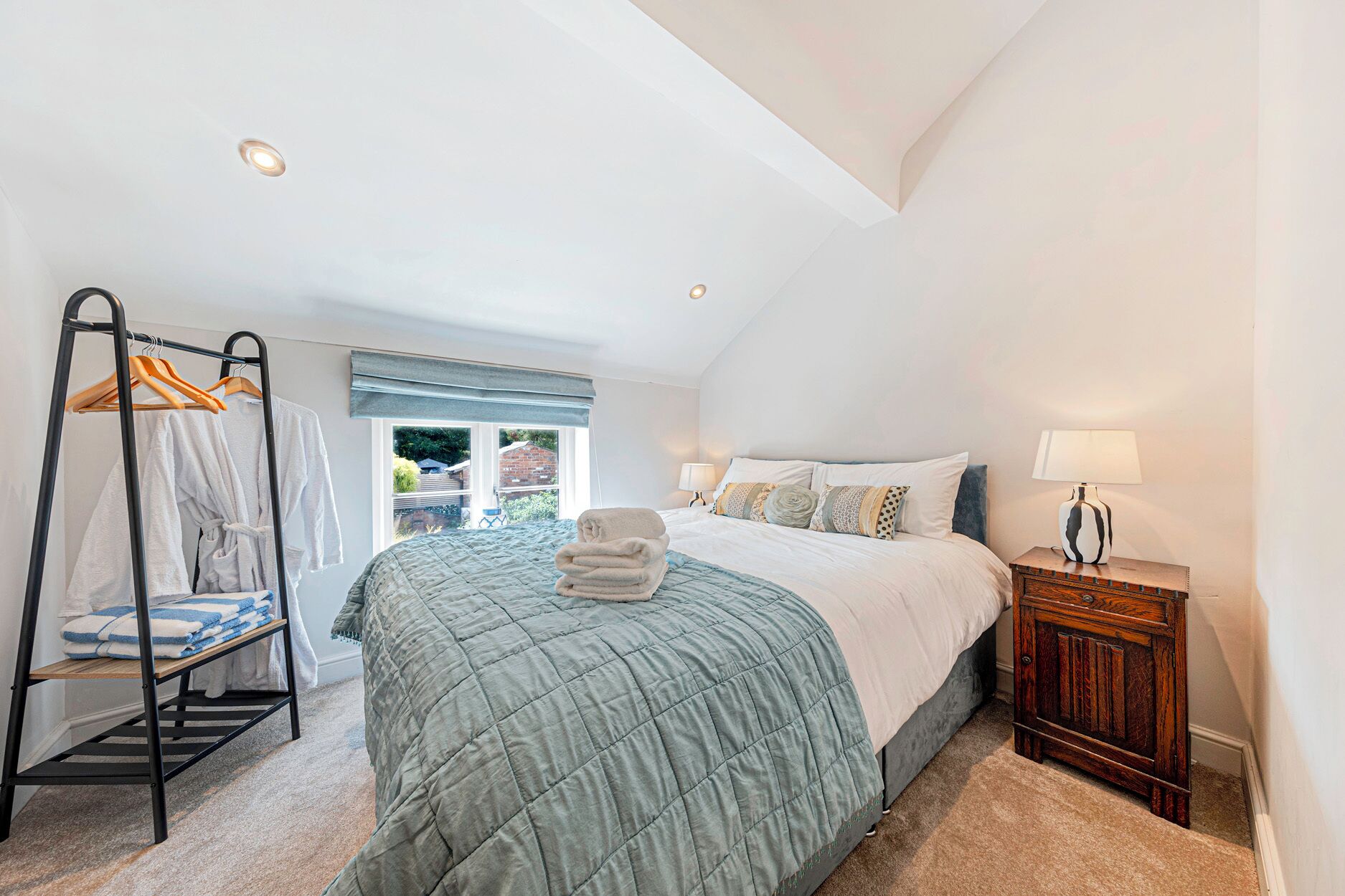 A bedroom with a bed, a bedside table with a lamp, a hanging clothes rack, and a window with a view of the outside.