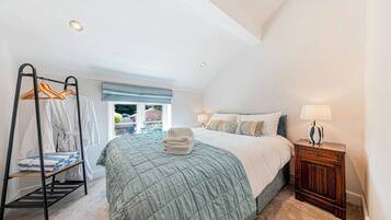 A bedroom with a bed, a bedside table with a lamp, a hanging clothes rack, and a window with a view of the outside.