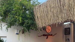 Un área de estar al aire libre con techo de paja, una mesa de madera y sillas. Hay plantas en macetas con flores en primer plano.