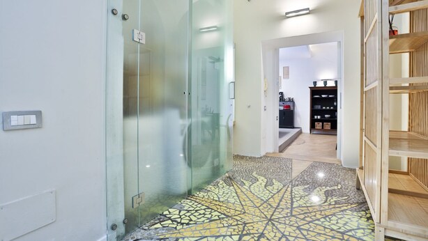 Um corredor com uma porta de vidro que dá para um banheiro, piso de azulejos e uma estante de madeira com objetos.