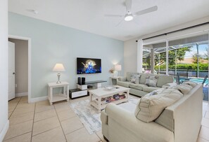 A living room with a flat-screen TV, a ceiling fan, a sofa, a coffee table, and a view of a pool through large windows. - Luxury Solterra Resort Retreat | Near Disney (Davenport)