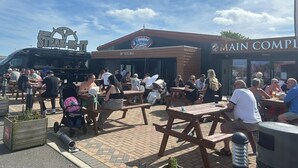 Ett utomhusområde med sittplatser, träbord och bänkar, en skylt för en sportbar och en parkeringsplats med en food truck.