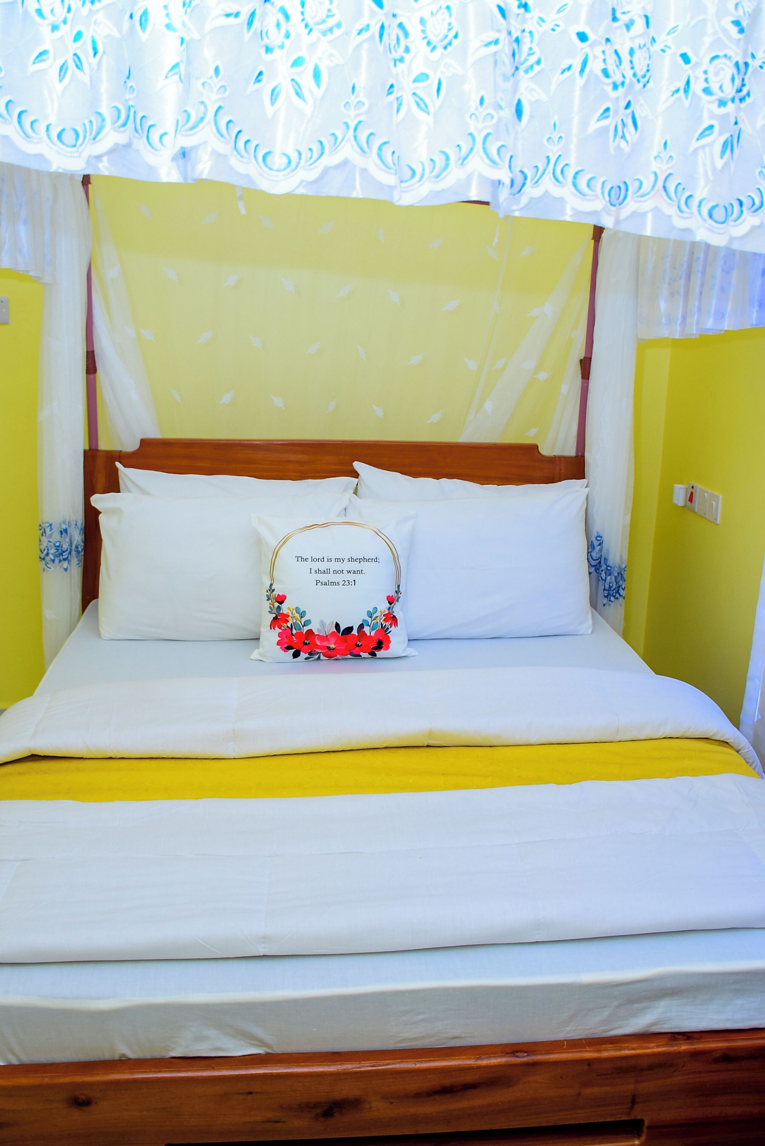A bed with a white mattress, a yellow bedspread, and a floral pillow with a Bible verse.