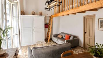 A room with a loft area, a grey sofa, a wooden dining table, and a staircase.