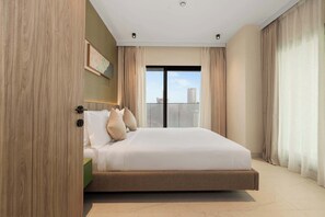 A modern bedroom with a large bed, a green nightstand, a painting on the wall, and a view of buildings through the window. - Executive Wellness Suite | Sauna, Pool & Gym (Jumeirah Village Triangle)