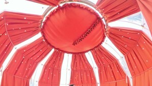 En person sitter på ett orange golv med ett geometriskt mönster, omgiven av röda draperier och en cirkelformad konstruktion med en öppning i mitten.