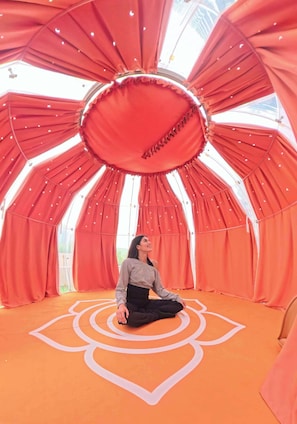 A person seated on an orange floor with a geometric pattern, surrounded by red curtains and a circular structure with a central opening. - Executive Wellness Suite | Sauna, Pool & Gym (Jumeirah Village Triangle)