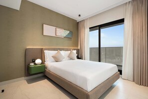 A modern hotel room with a large bed, a nightstand with a lamp, a painting on the wall, and a view of the city through the window. - Executive Wellness Suite | Sauna, Pool & Gym (Jumeirah Village Triangle)