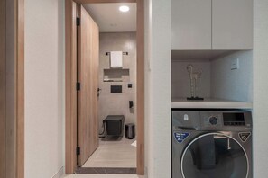 A modern laundry room with a front-loading washing machine, a trash bin, and a towel rack. - Tranquil Deluxe Stay with Pool, Gym & Wellness (Jumeirah Village Triangle)