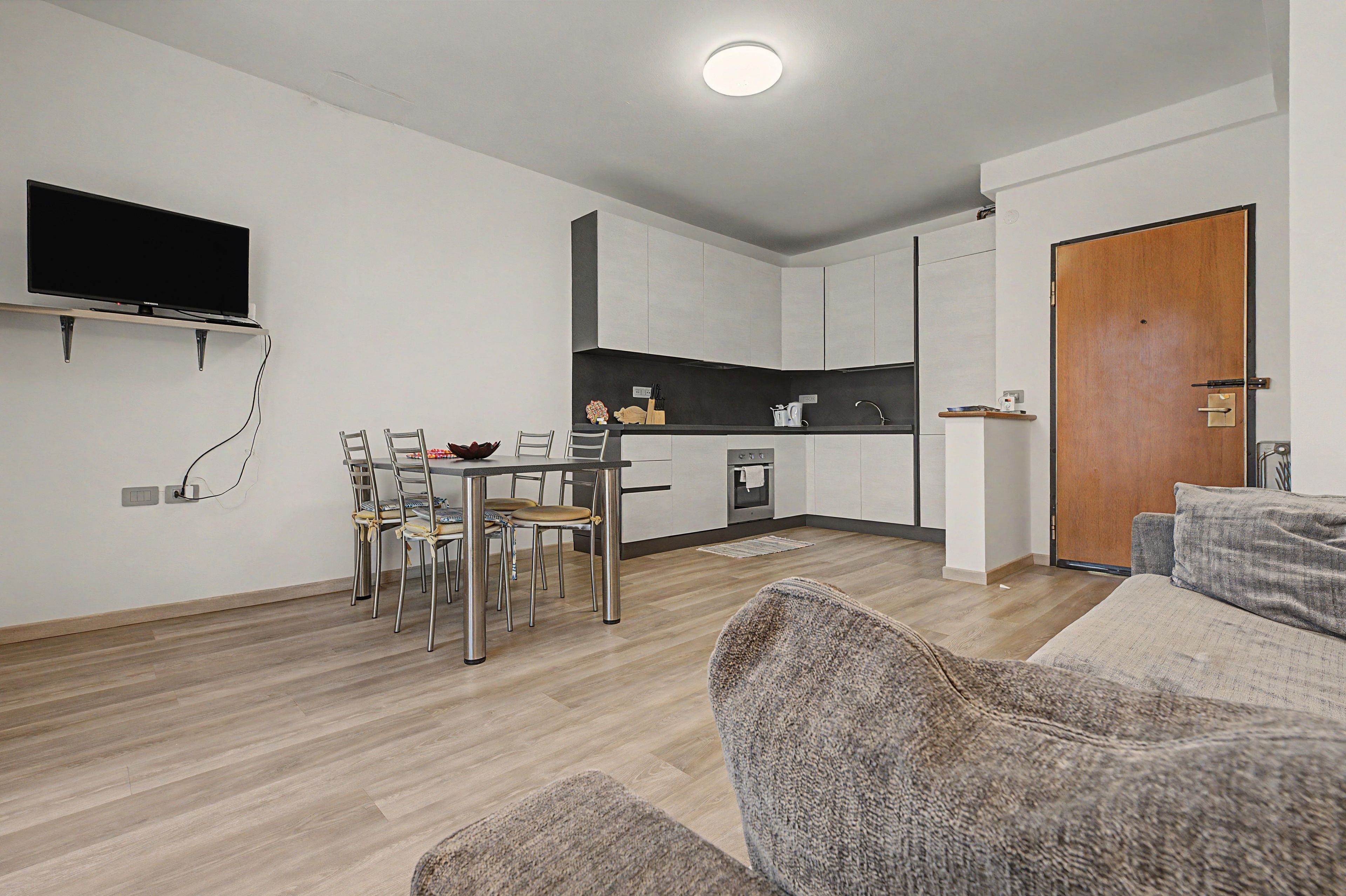 Une cuisine moderne avec des meubles blancs, un coin repas comprenant une table et des chaises, un téléviseur à écran plat fixé au mur et une porte en bois.