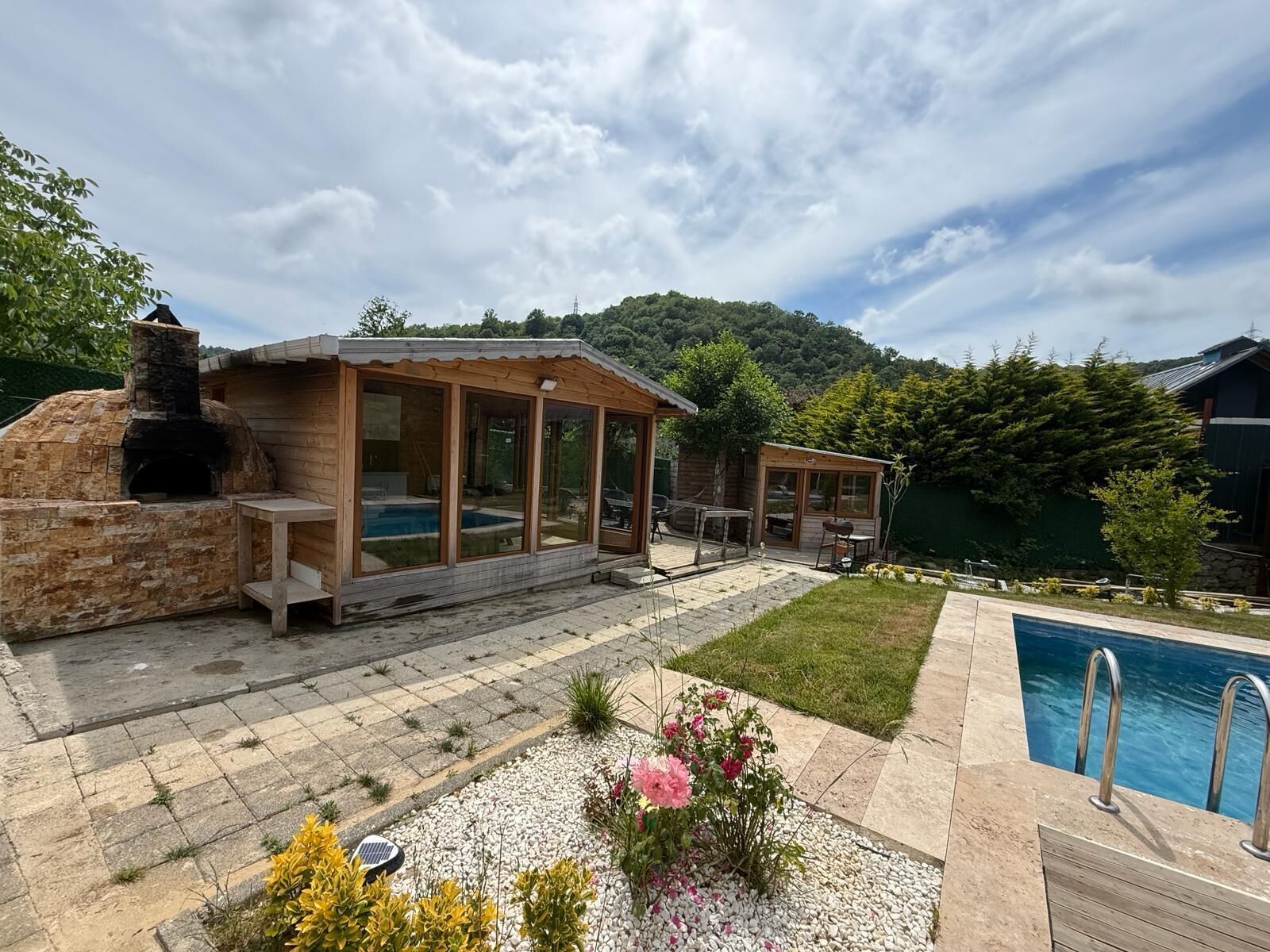 Cocina rústica al aire libre con horno de piedra, terraza de madera y piscina.