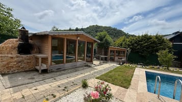 Cocina rústica al aire libre con horno de piedra, terraza de madera y piscina.
