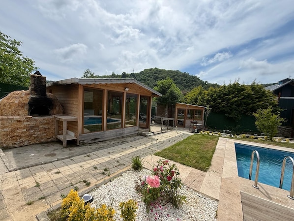 Cocina rústica al aire libre con horno de piedra, terraza de madera y piscina.