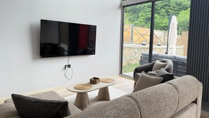 A modern living room with a flat-screen TV mounted on the wall, a stone fireplace, and a large window offering a view of the outdoors.