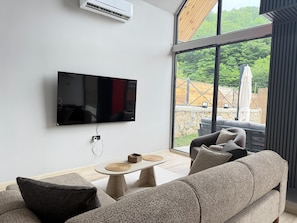 A modern living room with a flat-screen TV mounted on the wall, a stone fireplace, and a large window offering a view of the outdoors.