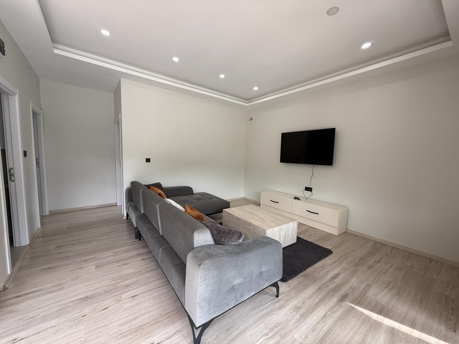 Una sala de estar moderna con un sofá seccional gris, una mesa de centro, un televisor de pantalla plana montado en la pared y un aparador blanco.