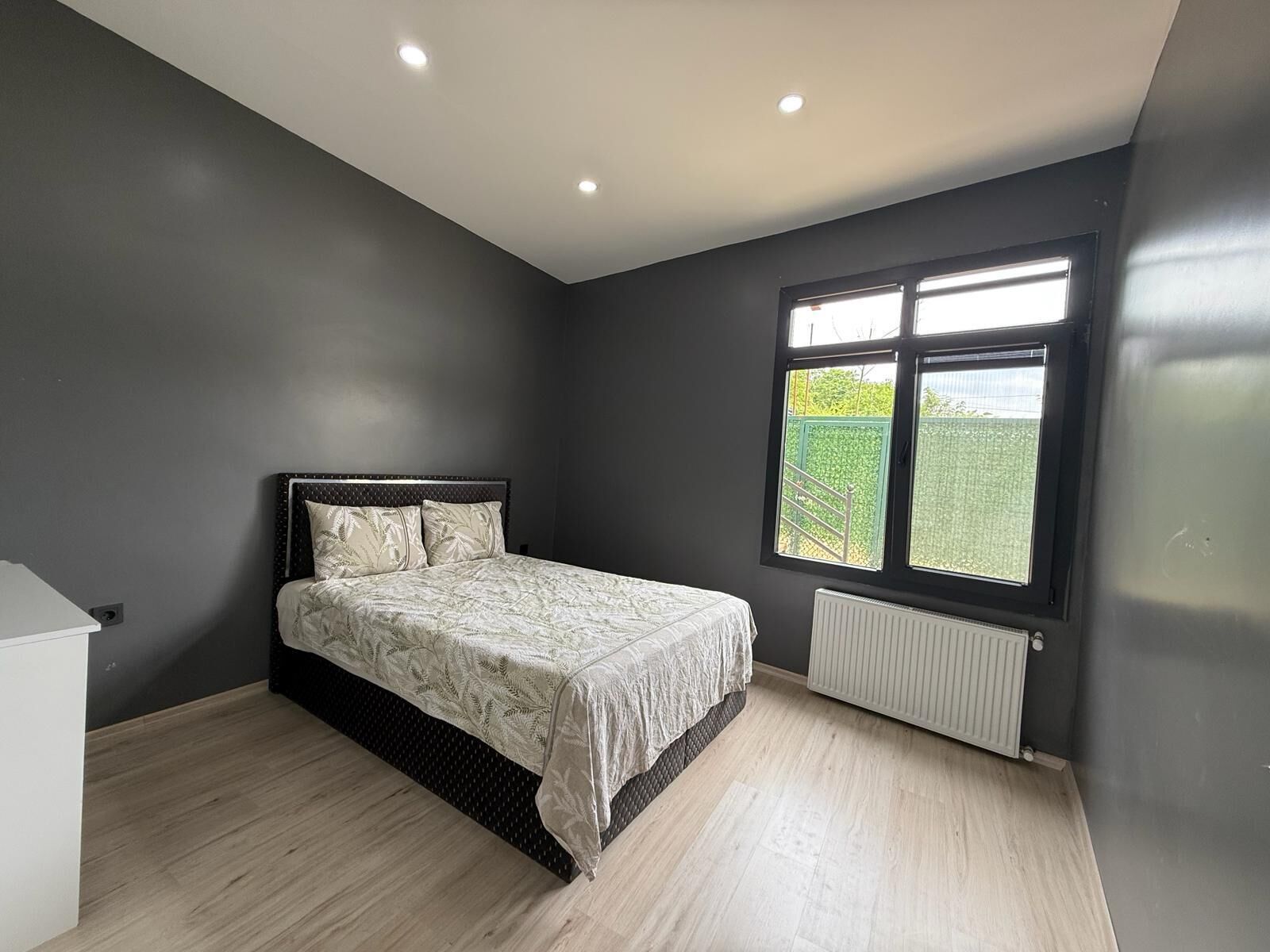 Un dormitorio con cama, un radiador y una ventana con vistas al verdor.
