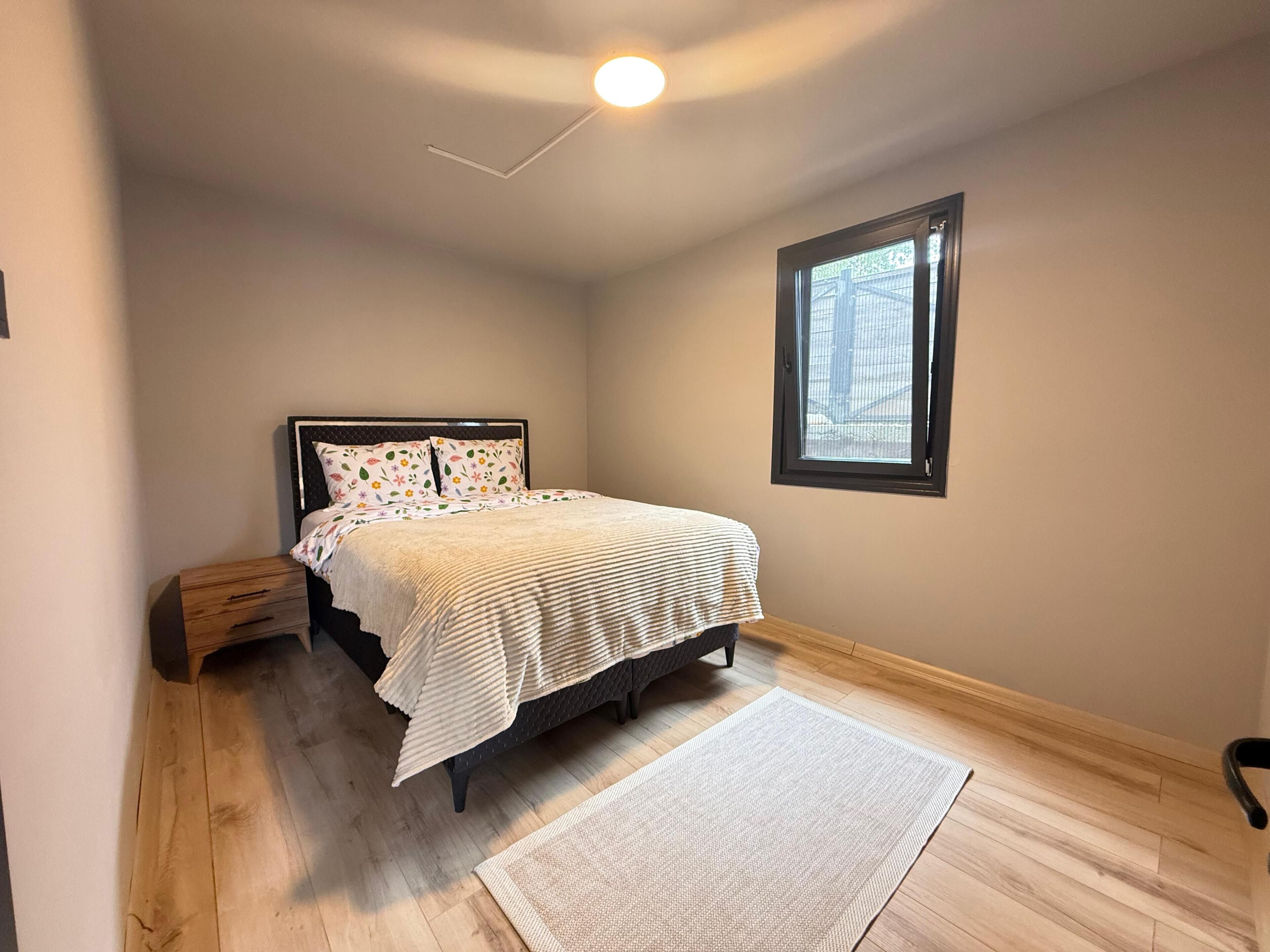Un dormitorio con cama, mesita de noche, ventana y luz de techo.