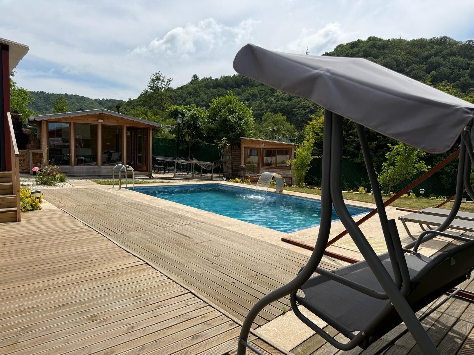 Una terraza de madera que conduce a una piscina con una cascada, rodeada de exuberante follaje y montañas al fondo.