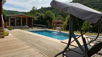 Una terraza de madera que conduce a una piscina con una cascada, rodeada de exuberante follaje y montañas al fondo.