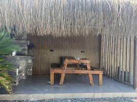 A wooden bench with a woven thatch roof, a stone planter with a palm plant, and a tiled floor.