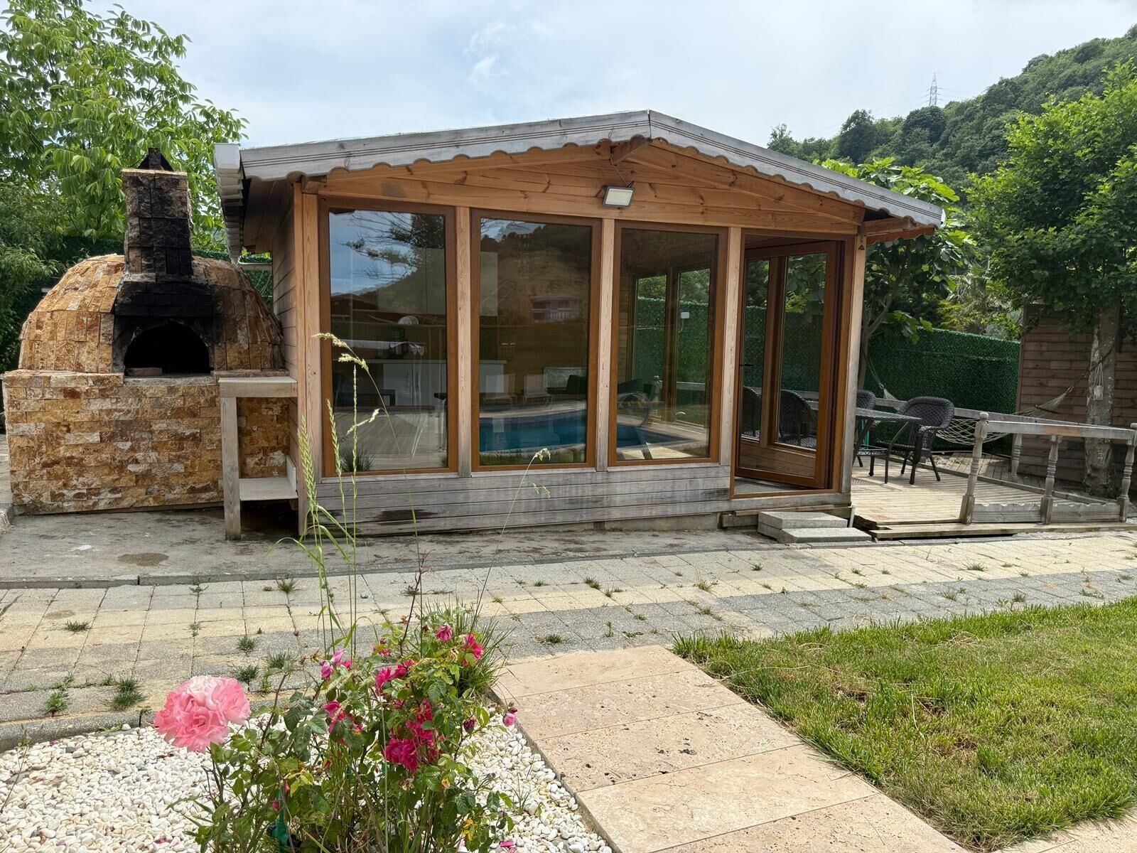 Una cocina exterior de ladrillo con chimenea, un porche cubierto de vidrio con piscina y un jardín con flores rosadas.