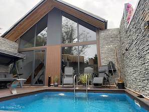 A modern outdoor pool area with a stone wall, lounge chairs, and a wooden deck.