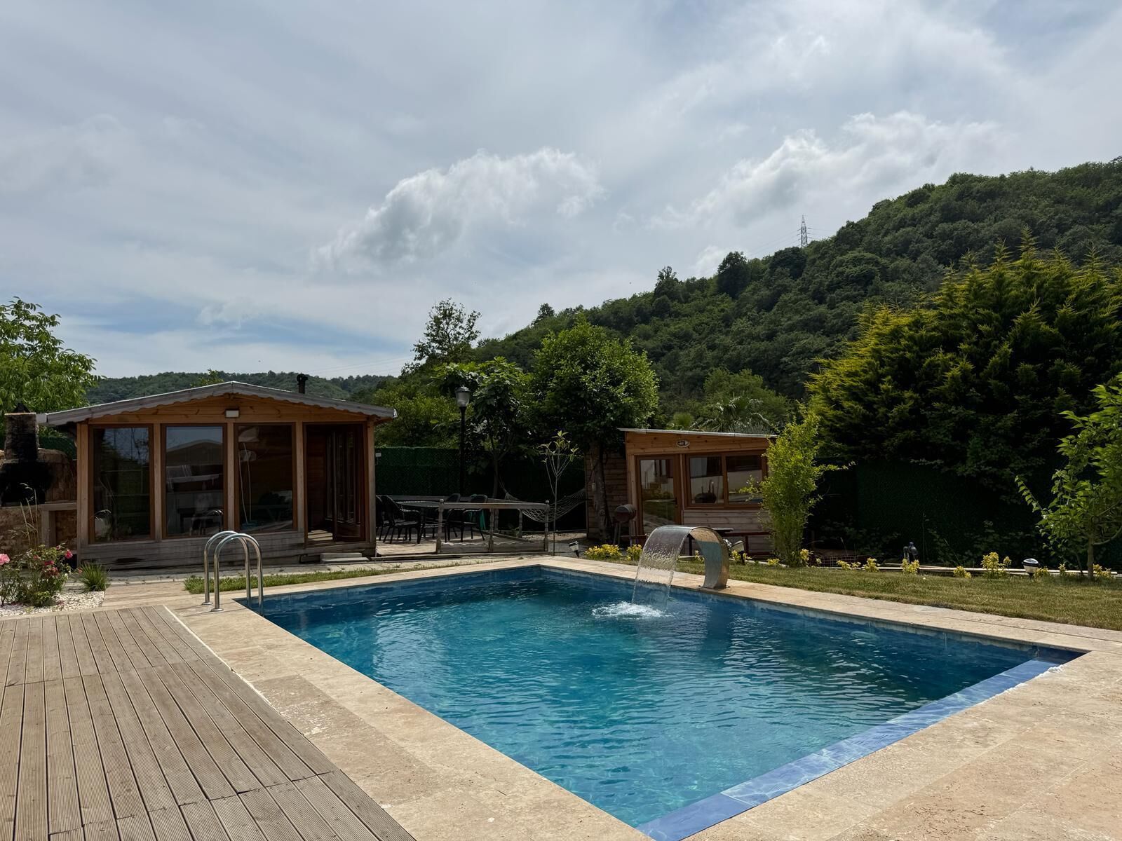 A swimming pool with a wooden deck, two wooden cabins, and a mountainous backdrop.
