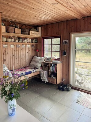 A cozy room with a wooden ceiling, a bed with a striped pillow, a shelf with baskets and jars, a window with a view of greenery, and a vase with flowers on a table.