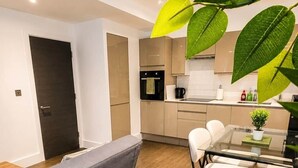 Una sala moderna con sofá, mesa de comedor y sillas, un área de cocina con armarios y electrodomésticos, y una puerta que da a otra habitación.