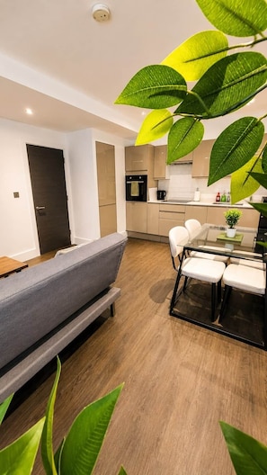 A modern living room with a sofa, dining table, and chairs, a kitchen area with cabinets and appliances, and a door leading to another room.