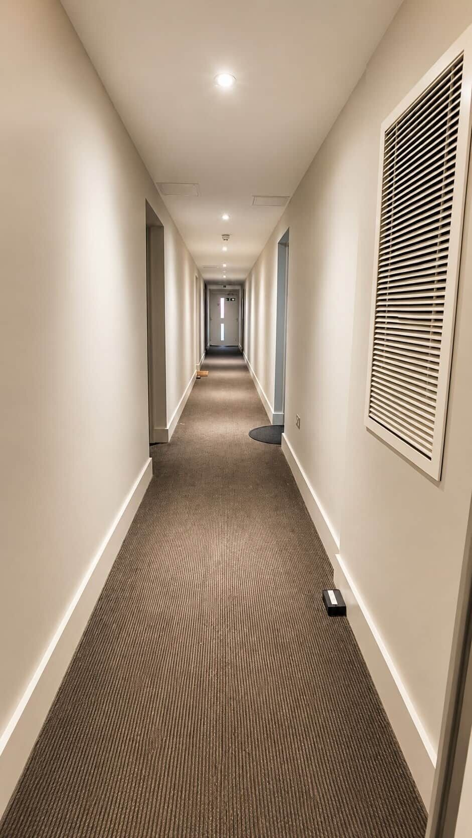 A hotel hallway with carpeted floors, a vent on the right, and a door at the end.