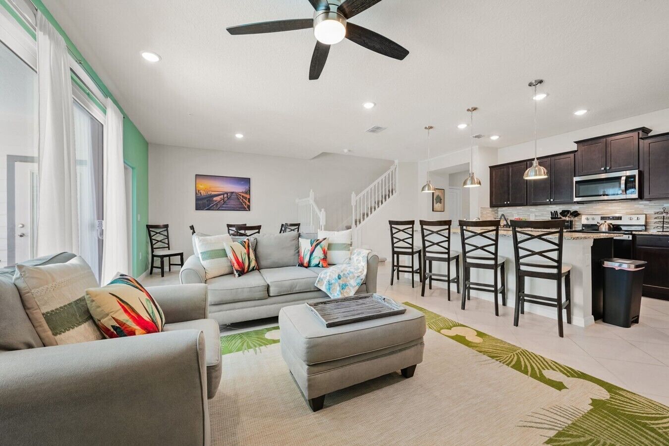 A modern living room with a grey sofa, a dining area with bar stools, a kitchen with dark cabinets, and a ceiling fan.
