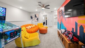 Una habitación con mesa de billar, sillones de mimbre, ventilador de techo y televisor montado en la pared.