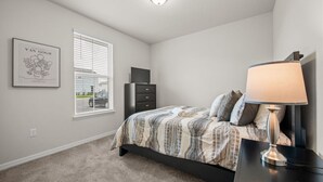 Un dormitorio con cama, mesita de noche, lámpara, cómoda, ventana con persianas y una obra de arte enmarcada en la pared.