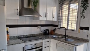 Een moderne keuken met witte kasten, een granieten aanrecht, een roestvrijstalen afzuigkap, een inbouwoven, een vaatwasser en een raam met jaloezieën.