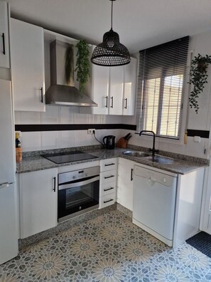 A modern kitchen with white cabinets, a granite countertop, a stainless steel range hood, a built-in oven, a dishwasher, and a window with blinds. - Yellowstone (AN)