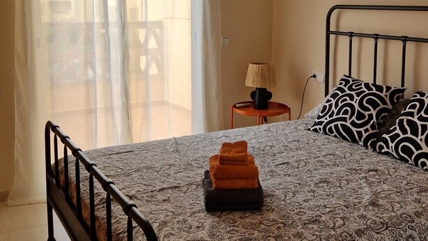 Een slaapkamer met een bed, een plafondventilator, een raam met gordijnen, een lamp en een schilderij aan de muur.