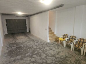 Ein Zimmer mit Betonboden, einer geschlossenen Tür und einer Treppe, die in eine höhere Ebene führt. Drei Stühle mit gemusterten Kissen sind vorhanden.