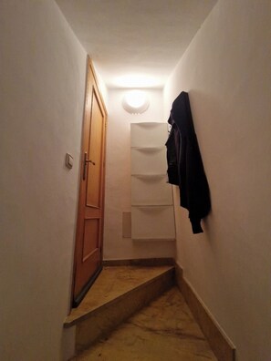A narrow hallway with a wooden door, a white wall-mounted shelf, and a black jacket hanging on the wall. - Yellowstone (AN)