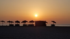 A beach scene with a row of thatched umbrellas and a small hut, likely a beach hut, with the sun setting in the background. - Yellowstone (AN)